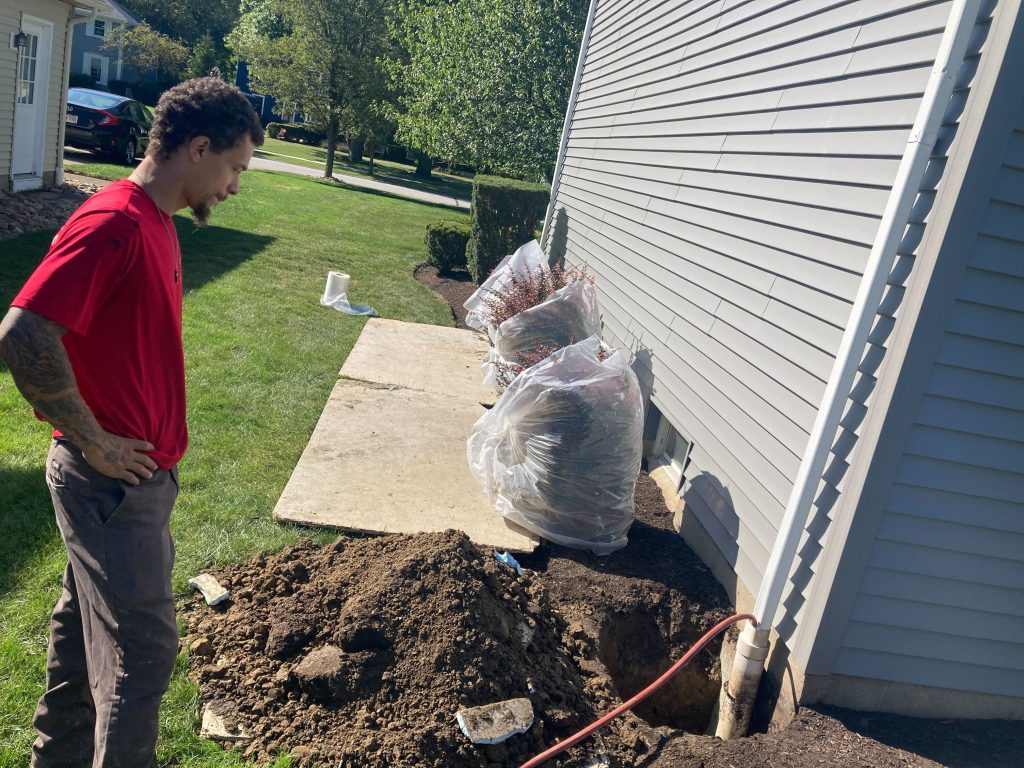 Man examining excavation site next to house for basement waterproofing, with dirt pile and covered plants visible.