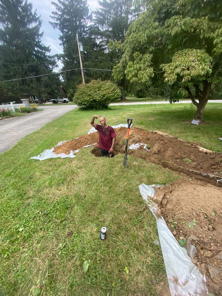 Man standing in a trench in a yard, holding a shovel, surrounded by dug soil and plastic sheeting, illustrating basement waterproofing preparation steps.