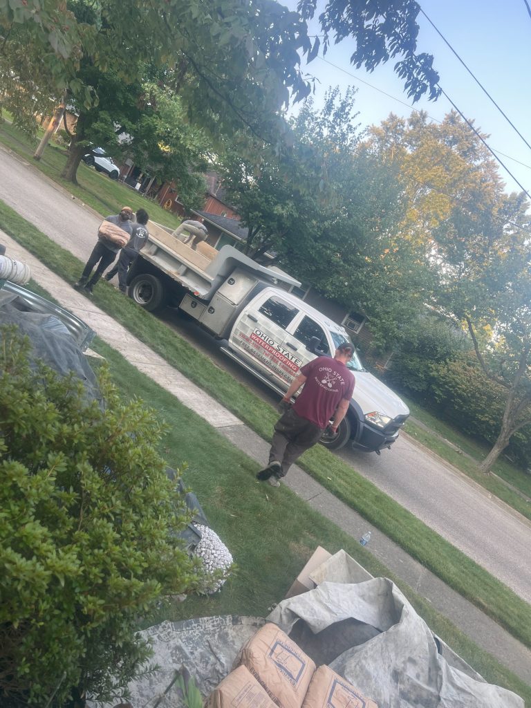 Workers from Ohio State Waterproofing unloading materials from a truck on a residential street, surrounded by landscaping and construction supplies.