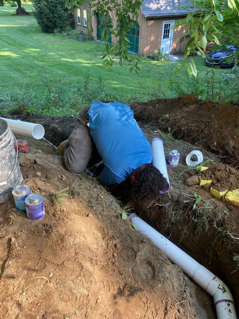 Person installing a perimeter drain system in a trench, surrounded by tools and pipes, in a residential yard setting.