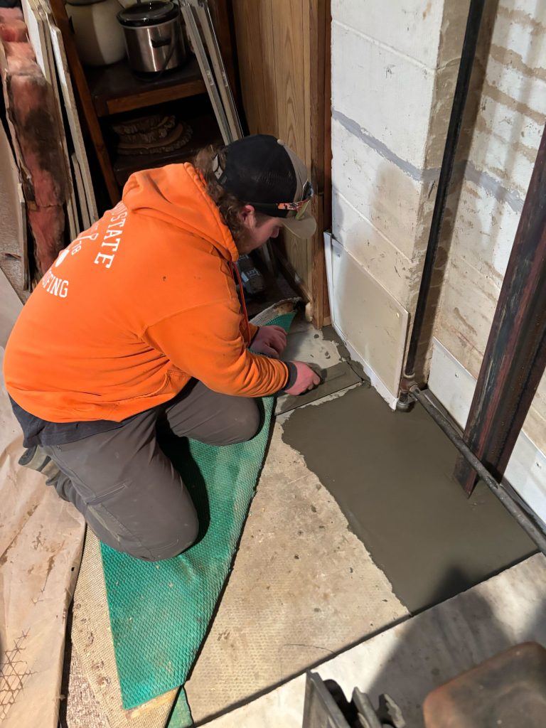 Worker applying concrete in a basement for waterproofing, wearing an orange Ohio State Waterproofing hoodie, with tools and materials visible in the background.