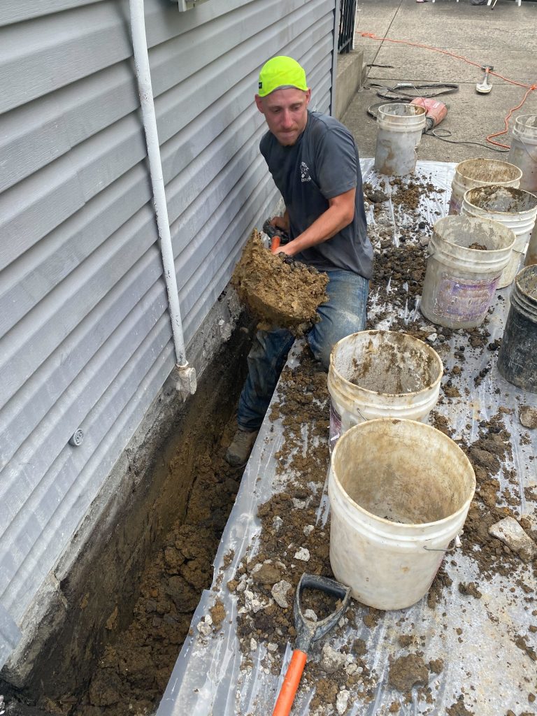 Worker excavating soil near a home's foundation for basement waterproofing, with buckets and tools visible, highlighting the process of foundation repair in Ohio.