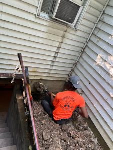 Worker in orange shirt performing foundation repair near basement entrance, emphasizing home waterproofing and prevention of water damage.