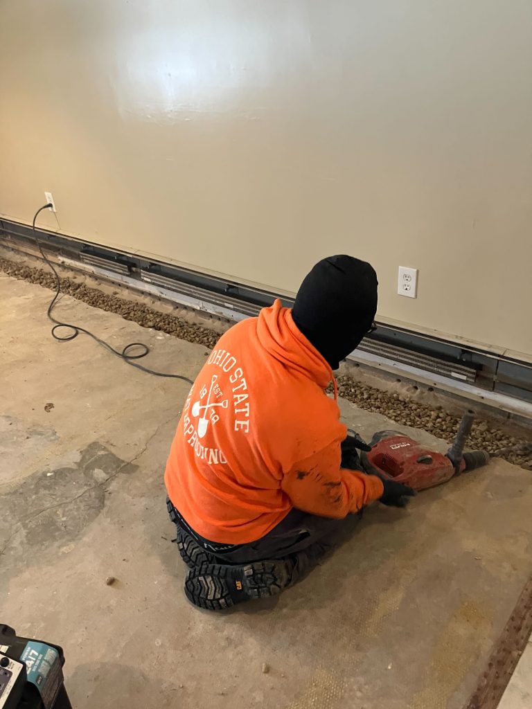 Worker in Ohio State Waterproofing sweatshirt using a power tool on a basement floor, addressing waterproofing issues.