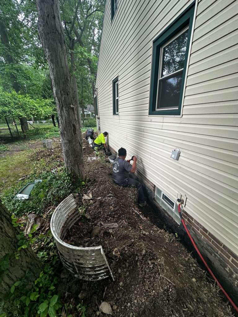 Workers installing waterproofing solutions on the exterior of a house, surrounded by dirt and landscaping, emphasizing basement protection from water damage.
