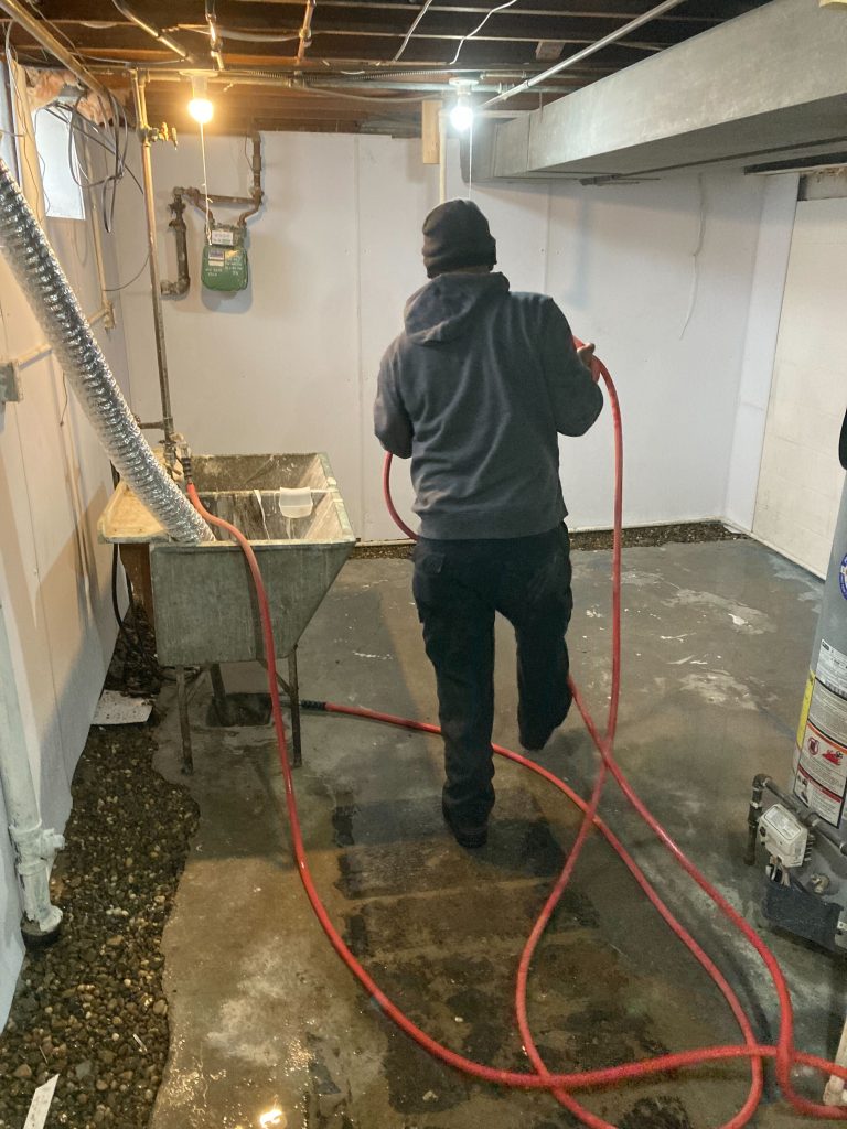 Person using a hose in a basement to manage water intrusion, with a utility sink and exposed plumbing visible, illustrating basement waterproofing efforts.
