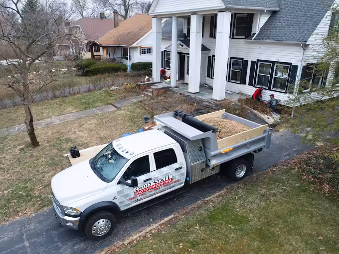 A white dump truck labeled "Ohio State Waterproofing" is parked on a driveway beside a large white house with columns. The scene is calm and overcast.