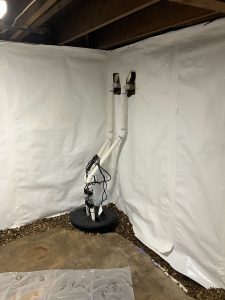 Basement corner with white waterproofing sheets, a sump pump system with vertical pipes, and visible wiring. Pebbles cover the floor around the pump, giving a clean, organized appearance.