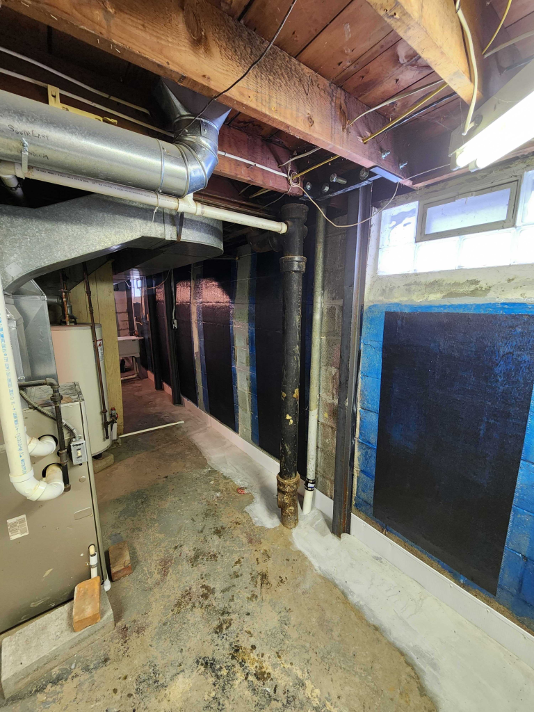 Basement with exposed wooden beams, ventilation ducts, and a black wall covering. Concrete floors show wear, and fluorescent lights create a dim, utilitarian feel.