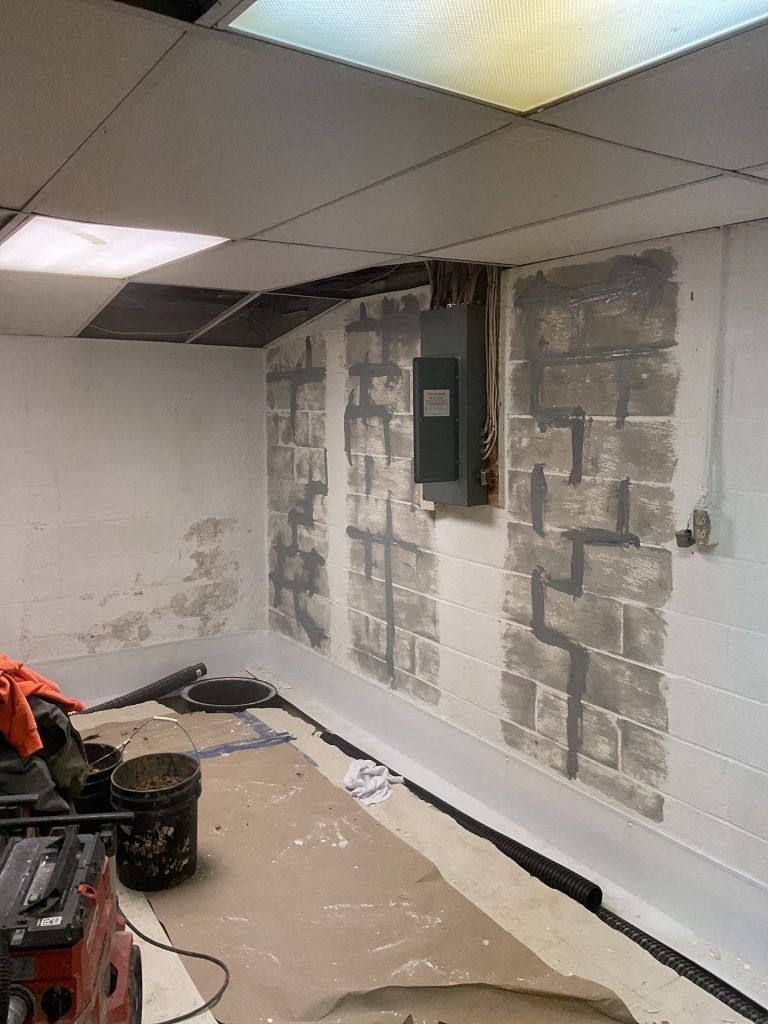 A partially renovated basement room with exposed cinder block walls, patched and sealed with gray lines. Tools, a bucket, and electrical panel are visible.