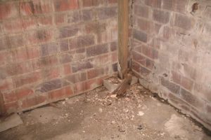 Corner of a basement with exposed brick walls, debris on the floor, and a wooden support beam, illustrating potential foundation issues relevant to basement waterproofing services.